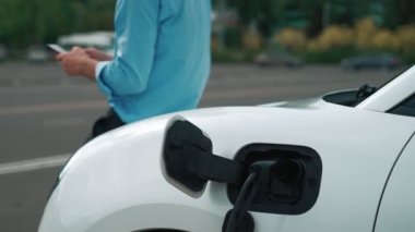 Takım elbiseli, hırslı bir işadamı elektrik kablosu ve yenilenebilir enerji enerjili bir elektrikli araçla şarj istasyonunda dururken elektrikli aracına yaslanıyor..