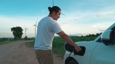 Elektrikli arabasıyla ilerici bir adam, EV arabası gelecekteki sürdürülebilir enerji kavramı olarak rüzgar türbiniyle yeşil alandaki enerji şarj etme istasyonundan enerji yüklüyor. Enerji üreteci olan elektrikli bir araç..