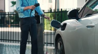 İlerici işadamı, şehir merkezinde dolaşmadan önce şarj cihazından elektrikli arabasına şarj prizi takıyor. Çevre dostu şarj edilebilir araba, sürdürülebilir ve temiz enerjiyle çalışıyor..