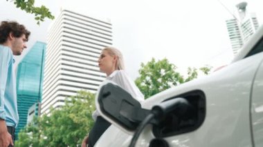 İlerici işadamı ve iş kadını, şehir merkezinde dolaşmadan önce şarj cihazını şarj istasyonundan elektrikli arabaya takıyor. Çevre dostu şarj edilebilir araba temiz enerjiyle çalışıyor..