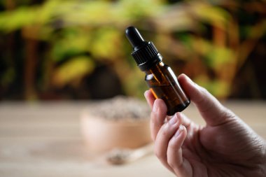 Eller, arka planda kenevir yaprağı olan bir şişe CBD yağı ve damlalık kapağını tutuyor. Tıbbi amaçlar için yasallaştırılmış CBD ürünü.