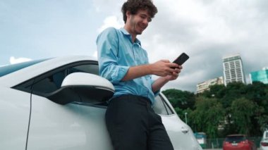 Takım elbiseli, hırslı bir işadamı elektrik kablosu ve yenilenebilir enerji enerjili bir elektrikli araçla şarj istasyonunda dururken elektrikli aracına yaslanıyor..