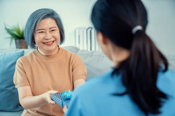 stock image A young caregiver hand over to her senior patient a blue gift box with blue ribbons at a contented living room.