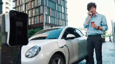 Takım elbiseli, hırslı bir işadamı elektrik kablosu ve yenilenebilir enerji enerjili bir elektrikli araçla şarj istasyonunda dururken elektrikli aracına yaslanıyor..