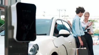 İlerici iş adamı ve iş kadını şehir merkezinde dolaşmadan önce şarj istasyonuna bağlı elektrikli arabaya yaslanıyor. Çevre dostu şarj edilebilir araba temiz enerjiyle çalışıyor..