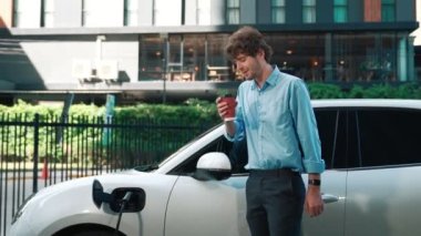 Takım elbiseli, hırslı bir işadamı elektrik kablosu ve yenilenebilir enerji enerjili bir elektrikli araçla şarj istasyonunda dururken elektrikli aracına yaslanıyor..