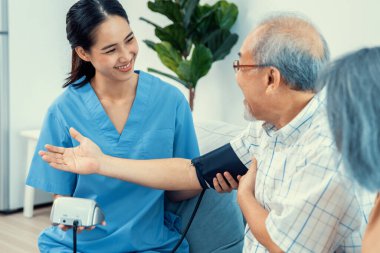 An elderly man having a blood pressure check by his personal caregiver with his wife sitting next to him in their home.