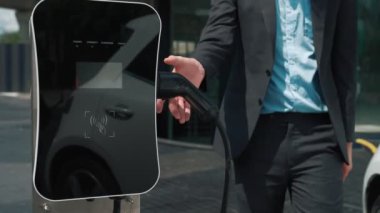 İlerici işadamı, şehir merkezinde dolaşmadan önce şarj cihazından elektrikli arabasına şarj prizi takıyor. Çevre dostu şarj edilebilir araba, sürdürülebilir ve temiz enerjiyle çalışıyor..