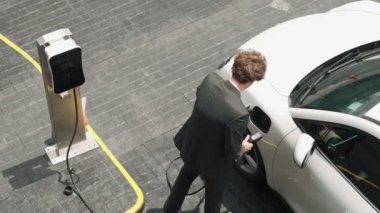 İlerici işadamı, şehir merkezinde dolaşmadan önce şarj cihazından elektrikli arabasına şarj prizi takıyor. Çevre dostu şarj edilebilir araba, sürdürülebilir ve temiz enerjiyle çalışıyor..