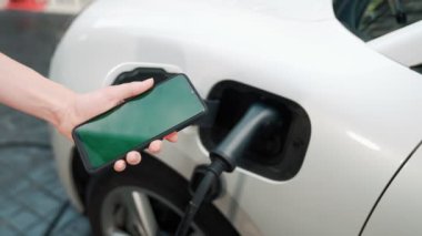 Taklit uzay için şarj istasyonuna bağlı elektrikli aracın yeşil ekran enerji durumunu gösteren bir model telefon. Temiz çevre için ilerici bir kavram. EV, yeşil yenilenebilir enerjiden güç alıyor.