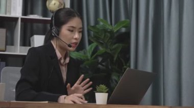 Kulaklık ve mikrofon takan iş kadını masasında çalışıyor ve dizüstü bilgisayar kullanıyor. Uzaktan kumandalı online toplantı için dizüstü bilgisayar kullanan hevesli kadın çalışanlar, ofiste iş videosu konferansı.