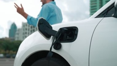 Takım elbiseli, hırslı bir işadamı elektrik kablosu ve yenilenebilir enerji enerjili bir elektrikli araçla şarj istasyonunda dururken elektrikli aracına yaslanıyor..
