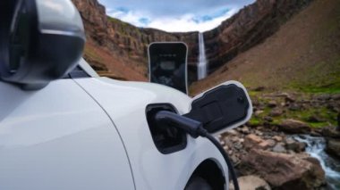Elektrikli araba, doğal ortam, şelale ve akış arkaplanındaki şarj istasyonundan enerji yüklüyor. Yenilenebilir enerji kaynaklı EV otomobillerin enerji sürdürülebilirliği kavramı.