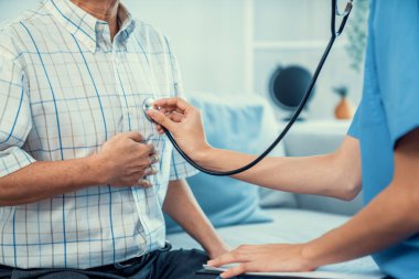 Caring young female doctor examining her contented senior patient with stethoscope in living room. Medical service for elderly, elderly sickness, declining health.