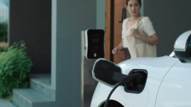 Evinde şarj edilen elektrikli bir araçla işten yeni dönen modern bir kadının ilerici yaşam tarzı. Elektrikli araç, sürdürülebilir temiz enerjiyle çalışıyor..