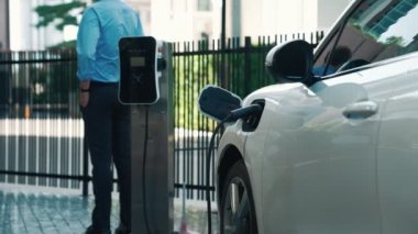 Takım elbiseli, hırslı bir işadamı elektrik kablosu ve yenilenebilir enerji enerjili bir elektrikli araçla şarj istasyonunda dururken elektrikli aracına yaslanıyor..