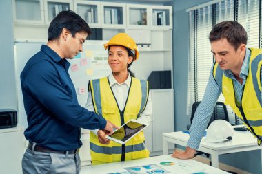 A team of competent engineers wearing safety equipment is working on blueprints with a tablet while also discussing with the investor in the office.