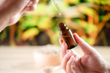 Hands holding a bottle of CBD oil and its dropper lid, with hemp leaf in the background. Legalized CBD product for medical purposes. clipart