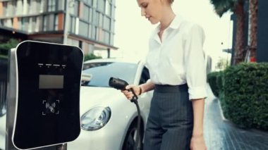 İlerici iş kadını şehir merkezinde dolaşmadan önce şarj cihazından elektrikli arabasına şarj prizi takıyor. Çevre dostu şarj edilebilir araba, sürdürülebilir ve temiz enerjiyle çalışıyor..