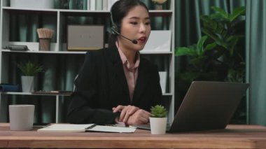 Kulaklık ve mikrofon takan iş kadını masasında çalışıyor ve dizüstü bilgisayar kullanıyor. Uzaktan kumandalı online toplantı için dizüstü bilgisayar kullanan hevesli kadın çalışanlar, ofiste iş videosu konferansı.