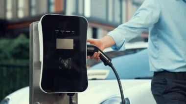 İlerici işadamı, şehir merkezinde dolaşmadan önce şarj cihazından elektrikli arabasına şarj prizi takıyor. Çevre dostu şarj edilebilir araba, sürdürülebilir ve temiz enerjiyle çalışıyor..