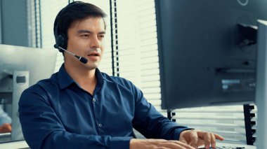 Young competent asian male call center agent working at his computer while simultaneously speaking with customers. Concept of an operator, customer service agent working in the office with headset. clipart