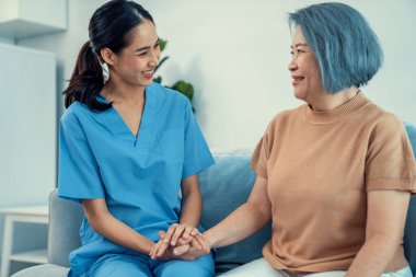 A caregiver rest her hands on the shoulders of a contented senior patient while she sitting on the sofa at home.