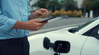 Takım elbiseli, hırslı bir işadamı elektrik kablosu ve yenilenebilir enerji enerjili bir elektrikli araçla şarj istasyonunda dururken elektrikli aracına yaslanıyor..