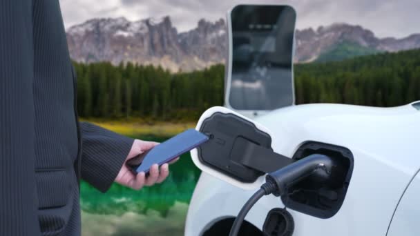 田舎の山を背景に充電ステーションでスマートフォンからEv車のバッテリー状態をチェックする進歩的なビジネスマン 現代の技術と自然のシーンの組み合わせ 環境に優しいEv車 — ストック動画