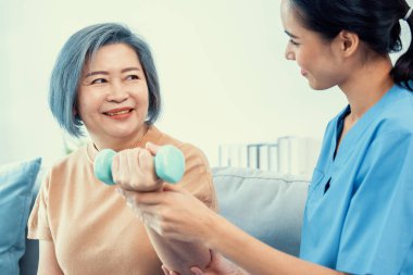 Caregiver helping contented senior woman exercise with dumbbell at home. Rehab for elder, senior medical care.