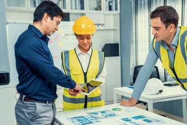A team of competent engineers wearing safety equipment is working on blueprints with a tablet while also discussing with the investor in the office.