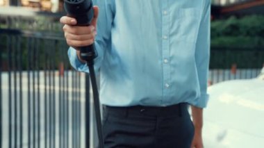 Takım elbiseli ve hırslı bir iş adamı kameraya güç kablosu takıyor, şehir merkezinde şarj oluyor. Elektrikli araç, yeşil çevre için yenilenebilir enerji kullanan EV araba.