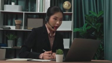 Kulaklık ve mikrofon takan iş kadını masasında çalışıyor ve dizüstü bilgisayar kullanıyor. Uzaktan kumandalı online toplantı için dizüstü bilgisayar kullanan hevesli kadın çalışanlar, ofiste iş videosu konferansı.