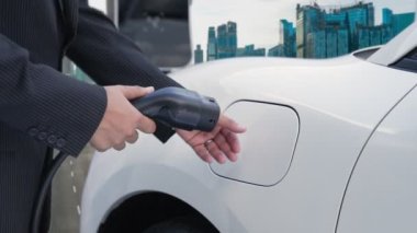 İlerici işadamı elektrikli aracını şarj kablosu ile şarj ediyor. Şarj istasyonundan şehrin arka planına bakıyor. Modern temiz enerji kampanyası konsepti için..