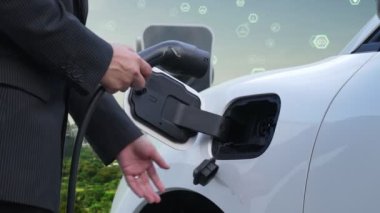 İlerleyen yeşil şehir ESG sembolü, elektrikli araçla birlikte. İş adamı EV arabalarını şarj etmek için şarj aletini şarj ediyor. Şehirdeki şarj istasyonunda ekolojik ve yeşil semboller var..
