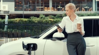 Şehir merkezinde dolaşmadan önce elektrikli araba ve şarj istasyonuna yaslanan ilerici iş kadını. Çevre dostu şarj edilebilir EV araba sürdürülebilir ve temiz enerjiyle çalışıyor.