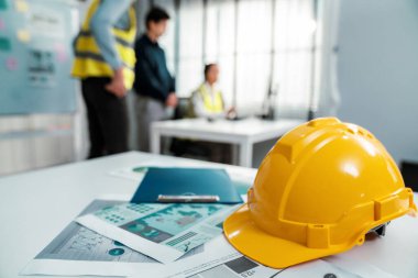 Concept of engineering equipments of protective helmet for competent engineer on piles of paper works. Blurred image of engineers and employer working together at the desk.