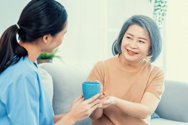 Female care taker serving her contented senior patient with a cup of coffee at home, smiling to each other. Medical care for pensioners, Home health care service.