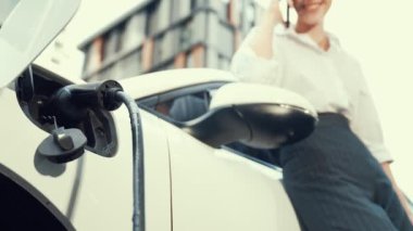 Şehir merkezinde dolaşmadan önce elektrikli araba ve şarj istasyonuna yaslanan ilerici iş kadını. Çevre dostu şarj edilebilir EV araba sürdürülebilir ve temiz enerjiyle çalışıyor.