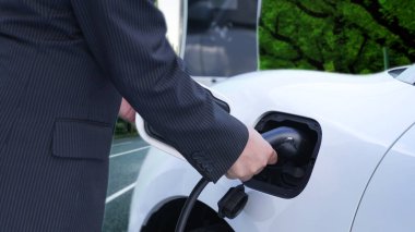 İlerici işadamı elektrikli aracını şarj kablosu ile şarj ediyor. Şarj istasyonundan şehrin arka planına bakıyor. Modern temiz enerji kampanyası konsepti için..