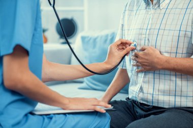 Caring young female doctor examining her contented senior patient with stethoscope in living room. Medical service for elderly, elderly sickness, declining health.
