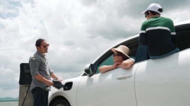 İlerici mutlu aile kavramı elektrikli araçla rüzgar çiftliğinde eğleniyor. Rüzgar türbini jeneratöründen gelen temiz yenilenebilir enerji ile şarj edilen elektrikli araç..