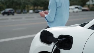 Takım elbiseli, hırslı bir işadamı elektrik kablosu ve yenilenebilir enerji enerjili bir elektrikli araçla şarj istasyonunda dururken elektrikli aracına yaslanıyor..