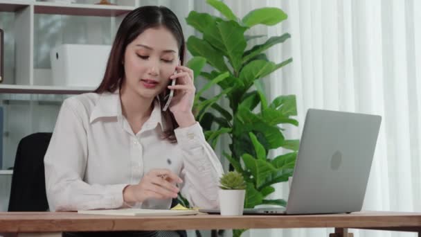 若い熱狂的な実業家は ビジネスコールで話をし ノートパソコンで彼女の机にメモを書きます 女性従業員が電話で話をし オフィスで紙にビジネスタスクを書く — ストック動画