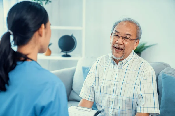 Stock image Caring young female doctor examining her contented senior patient with stethoscope in living room. Medical service for elderly, elderly sickness, declining health.