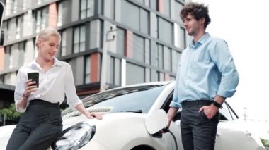İlerici iş adamı ve iş kadını şehir merkezinde dolaşmadan önce şarj istasyonuna bağlı elektrikli arabaya yaslanıyor. Çevre dostu şarj edilebilir araba temiz enerjiyle çalışıyor..