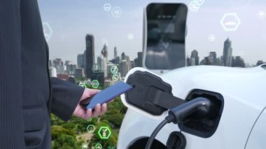 İlerleyen yeşil şehir ESG sembolü, elektrikli araçla birlikte. İş adamı EV arabalarını şarj etmek için şarj aletini şarj ediyor. Şehirdeki şarj istasyonunda ekolojik ve yeşil semboller var..