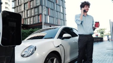 Takım elbiseli, hırslı bir işadamı elektrik kablosu ve yenilenebilir enerji enerjili bir elektrikli araçla şarj istasyonunda dururken elektrikli aracına yaslanıyor..