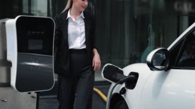 İlerici iş kadını, şehir merkezinde dolaşmadan önce şarj cihazının fişini şarj istasyonundan elektrikli arabasına çekti. Çevre dostu şarj edilebilir araba, sürdürülebilir ve temiz enerjiyle çalışıyor..