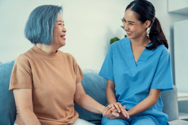 A caregiver rest her hands on the shoulders of a contented senior patient while she sitting on the sofa at home.
